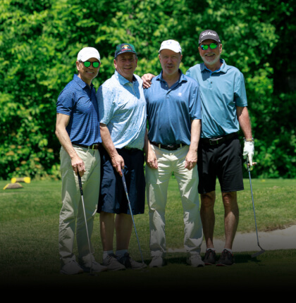 Four men stand on a sunny golf course holding clubs, smiling warmly. They wear golf attire and caps, with lush green trees in the background.