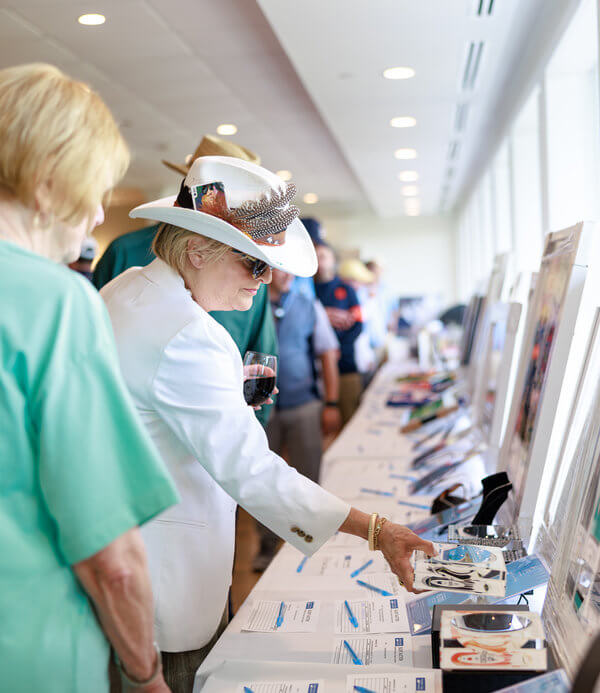A woman in a white hat and blazer examines auction items on a long table, holding a glass of red wine. The atmosphere is lively and social.