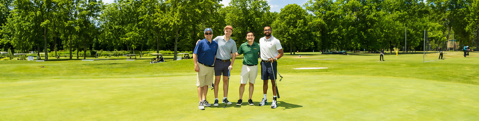 Group of four men posing on a golf course, each holding a golf club, under a clear blue sky