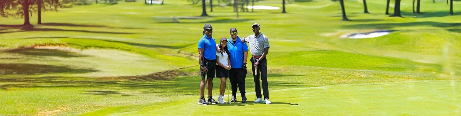 A group of four golfers stand together on a sunny green golf course, smiling and holding their clubs, dressed in sporty attire